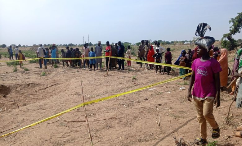 A group of people stands behind yellow caution tape on a sandy field under a clear sky, with sparse vegetation in the background.