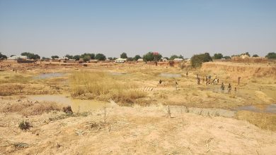 Dry riverbed with children playing, surrounded by mud and sparse vegetation; village huts in the background under a clear blue sky.