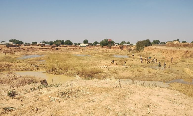 Dry riverbed with children playing, surrounded by mud and sparse vegetation; village huts in the background under a clear blue sky.