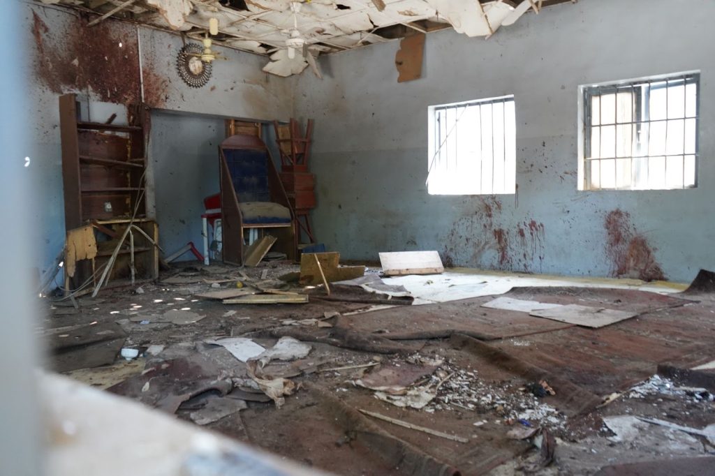 Dilapidated room with debris, peeling ceiling, and stained walls. Sunlight filters through barred windows.