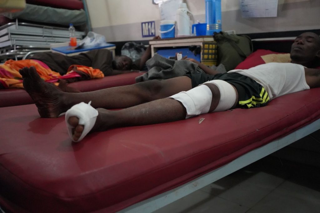 A person lies on a hospital bed with a bandaged knee and foot. Another person is resting on a bed in the background.