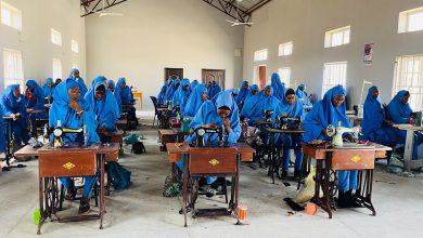 Women in blue hijabs sewing in a classroom, focusing on their tasks, surrounded by sewing machines and materials.