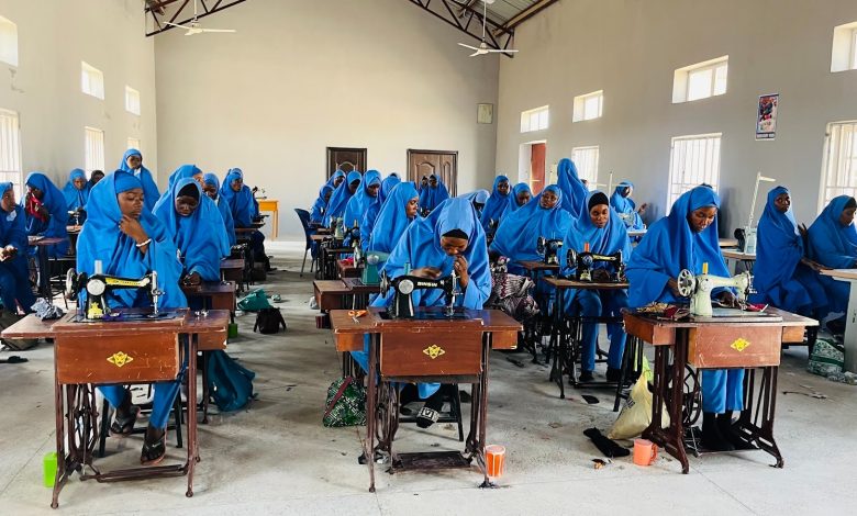 Women in blue hijabs sewing in a classroom, focusing on their tasks, surrounded by sewing machines and materials.