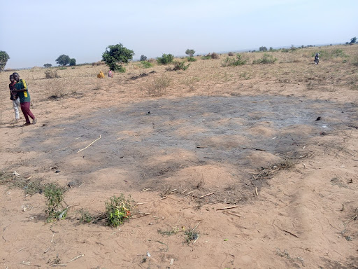 A barren field with a large patch of dry, cracked earth. A few people are walking in the distance, with scattered bushes and trees around.