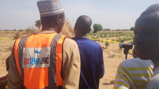 People gathered in an open field, one wearing a bright safety vest with text, under a clear sky.