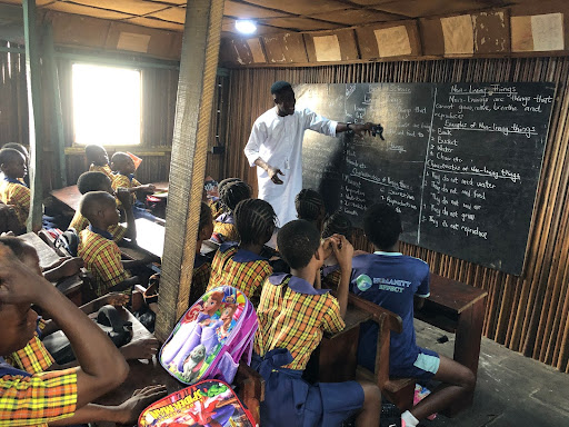 Teacher instructing students in a classroom with wooden walls and a chalkboard. Children are seated in uniforms, observing and taking notes.