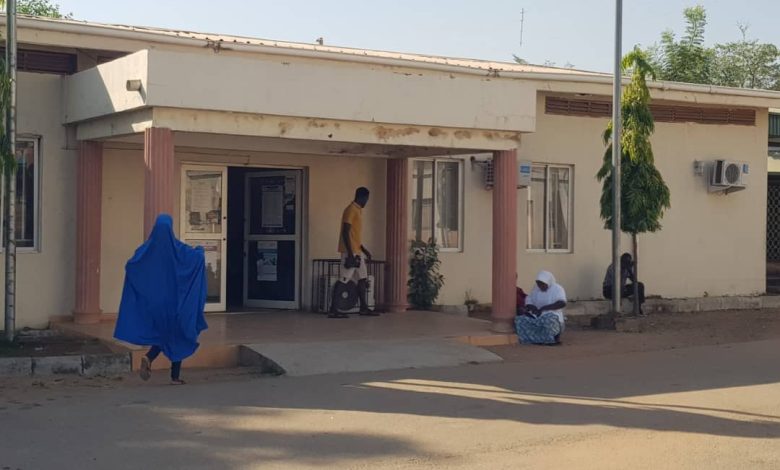 A person in blue clothing walks toward a small building; another sits nearby. Trees and clear sky in the background.