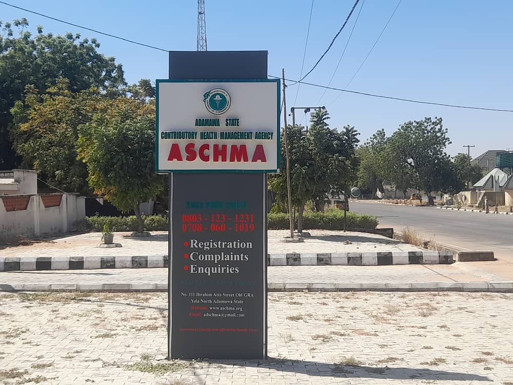 Sign for Adamawa State Contributory Health Management Agency (ASCHMA) with contact info and services, located on a paved roadside.