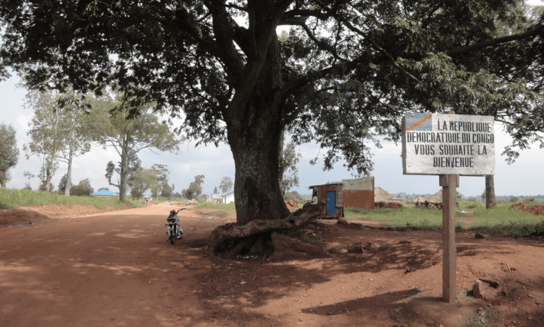 Dirt road with a large tree and a sign saying "La République Démocratique du Congo vous souhaite la bienvenue" on the right.