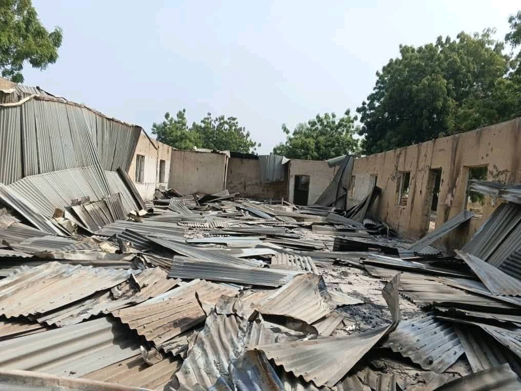 Collapsed building with a corrugated metal roof scattered on the ground, surrounded by damaged walls and trees in the background.
