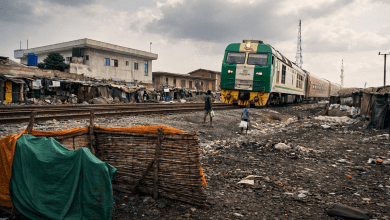 Green train passing through a rundown area with makeshift houses, as two people walk beside the tracks under a cloudy sky.