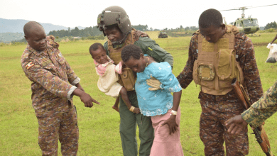 Soldiers assist a man carrying a child and escort a woman on a grassy field with helicopters in the background.