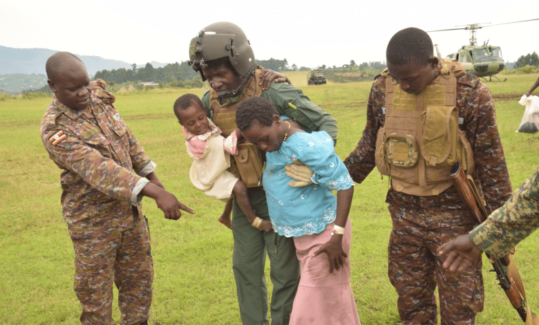 Soldiers assist a man carrying a child and escort a woman on a grassy field with helicopters in the background.