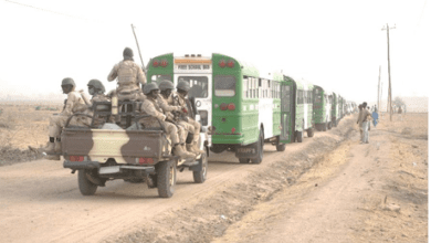 Military personnel on a jeep escort a convoy of green buses along a dirt road in a rural area.