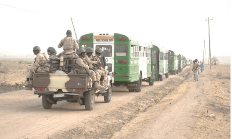 Military personnel on a jeep escort a convoy of green buses along a dirt road in a rural area.