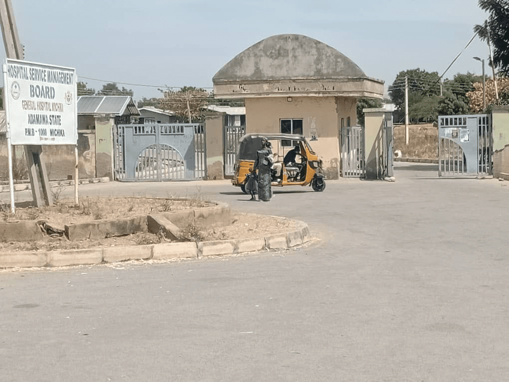 A person in a yellow auto rickshaw outside a hospital gate in Adamawa State, Nigeria, next to a sign for the General Hospital Michika.