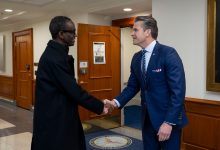 Two men in formal attire shaking hands in a hallway with wood paneling.
