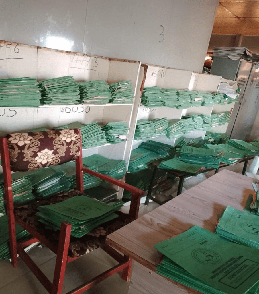 Room filled with stacks of green folders on shelves, a chair, and a table, suggesting a busy office environment.