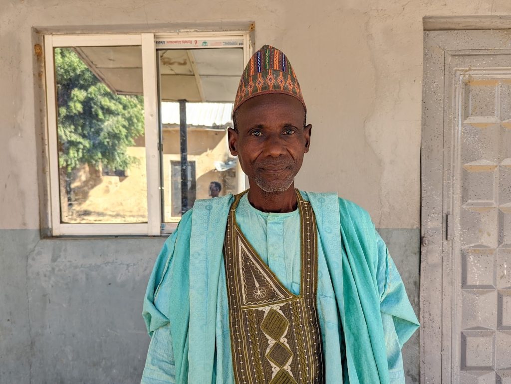 Elderly man in traditional attire stands in front of a window and door, with an expression of calmness.