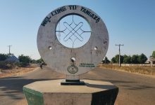 Welcome sign in Tangaza, Nigeria, with a circular design in the center. Road, trees, and buildings are visible in the background.