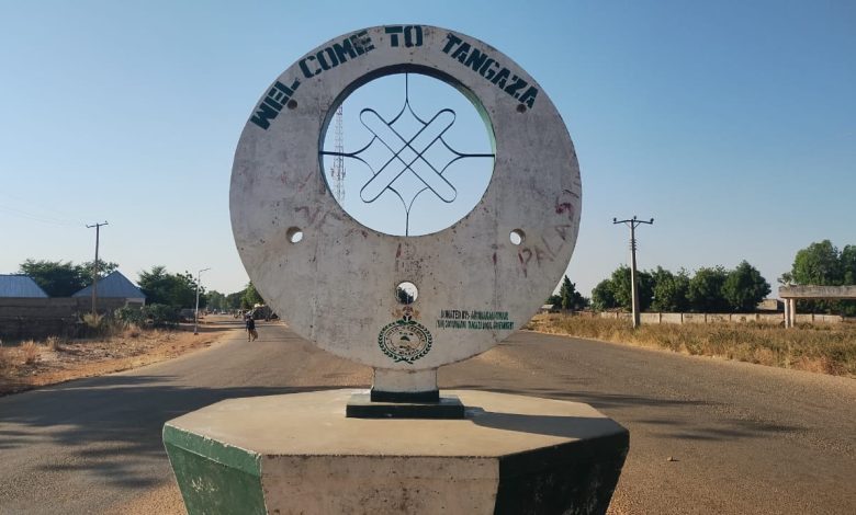 Welcome sign in Tangaza, Nigeria, with a circular design in the center. Road, trees, and buildings are visible in the background.