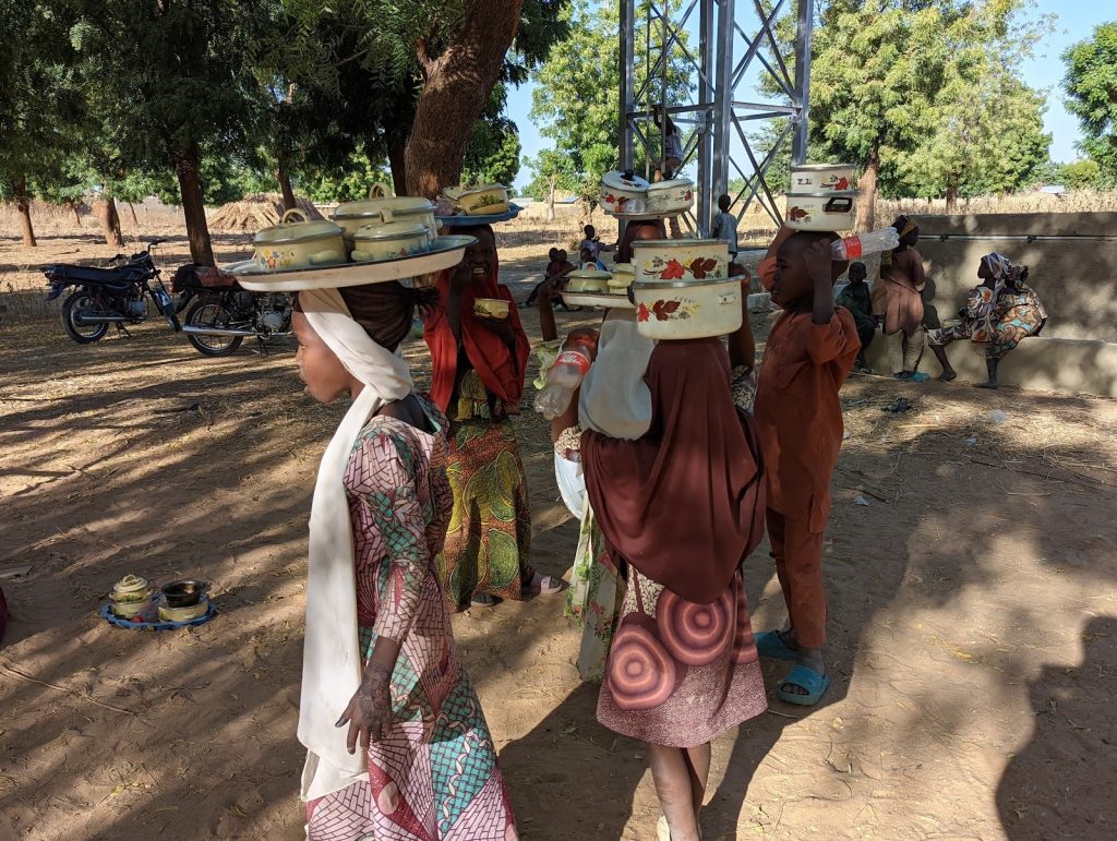 Children balancing trays of pots on their heads under trees, with motorcycles and others sitting nearby.