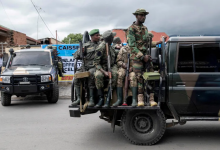 Soldiers in camouflage gear sitting and standing on a military vehicle parked on a street, with another vehicle in the background.