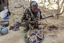 Person preparing poultry outdoors with traditional attire, sitting on the ground next to a stone wall.