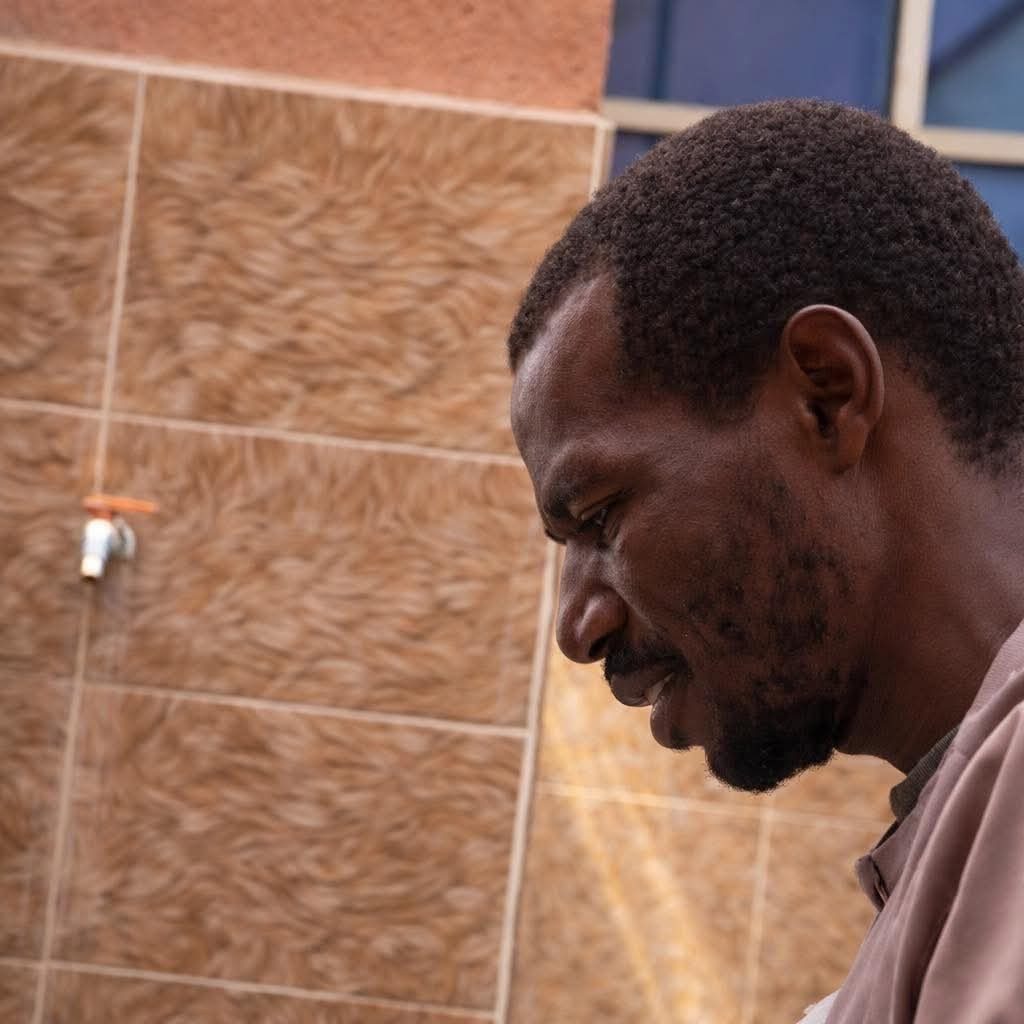 A person looking down, standing in front of a textured tiled wall with a faucet in the background.