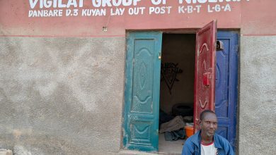 Man sitting outside a building with a sign reading "Vigilat Group of Nigeria" above blue and red open doors.