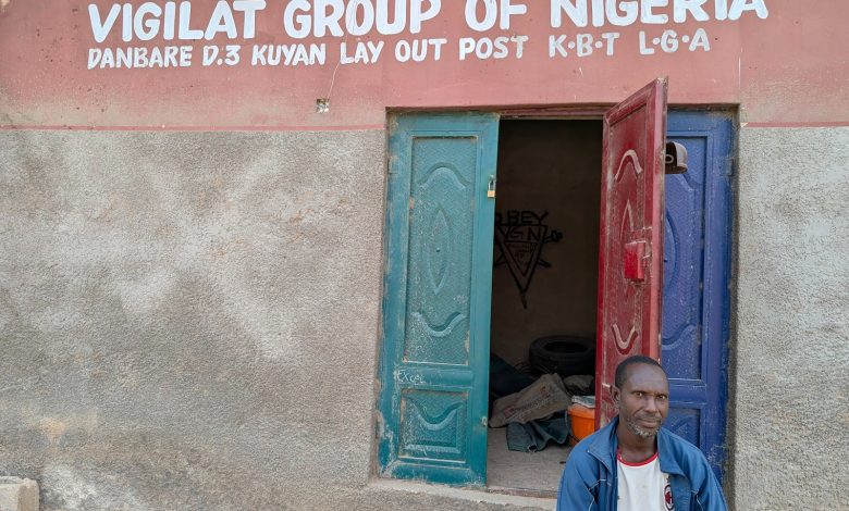 Man sitting outside a building with a sign reading "Vigilat Group of Nigeria" above blue and red open doors.