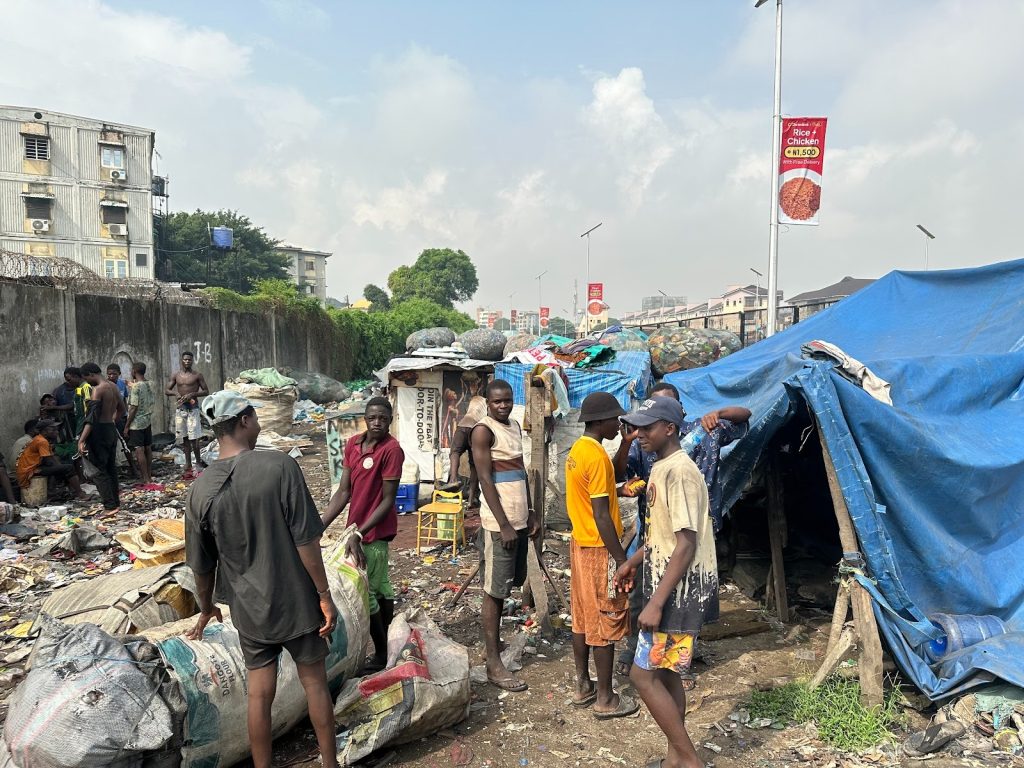 People gather in an area with scattered trash and makeshift shelters, engaged in various activities under a partly cloudy sky.