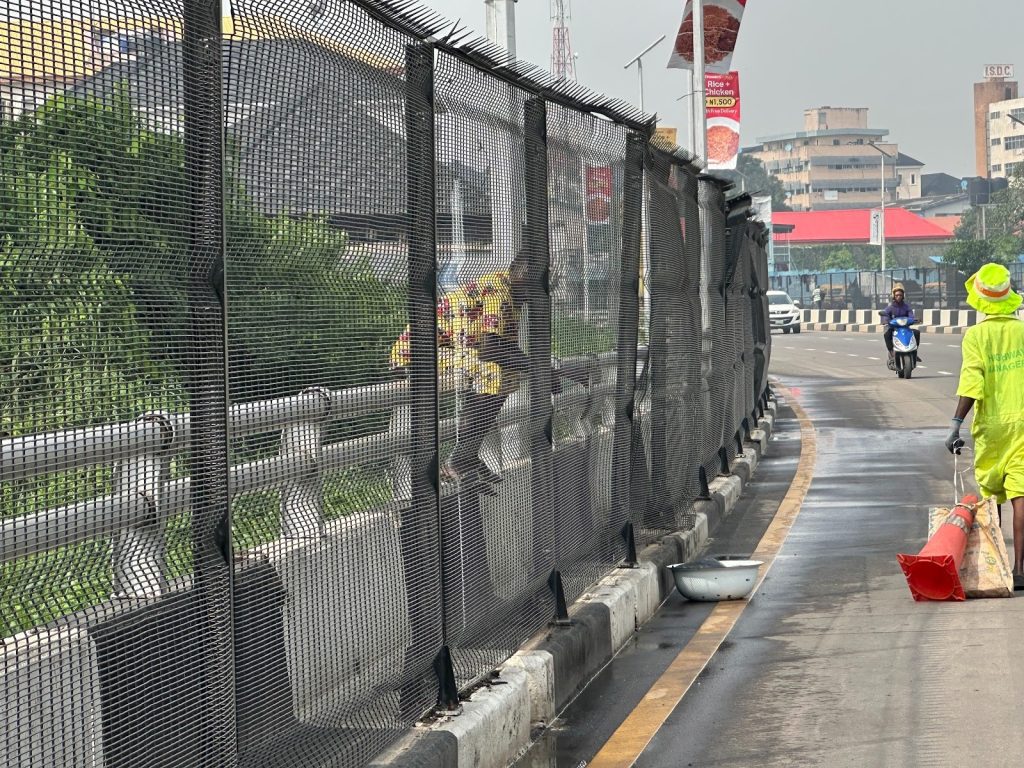 A worker in a neon outfit pulls a cart along a wet urban road with a mesh fence. A motorcyclist rides on the opposite side.
