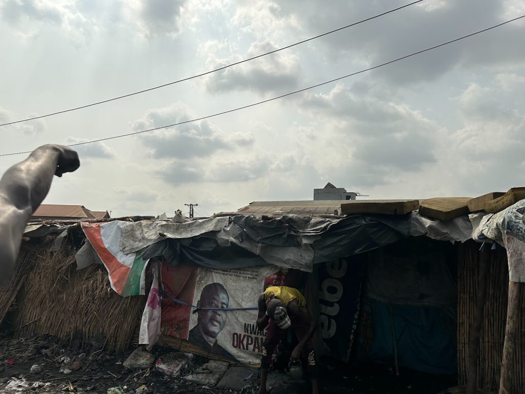 A person bends over outside a makeshift structure with posters on it, under a cloudy sky, with an outstretched arm in the foreground.
