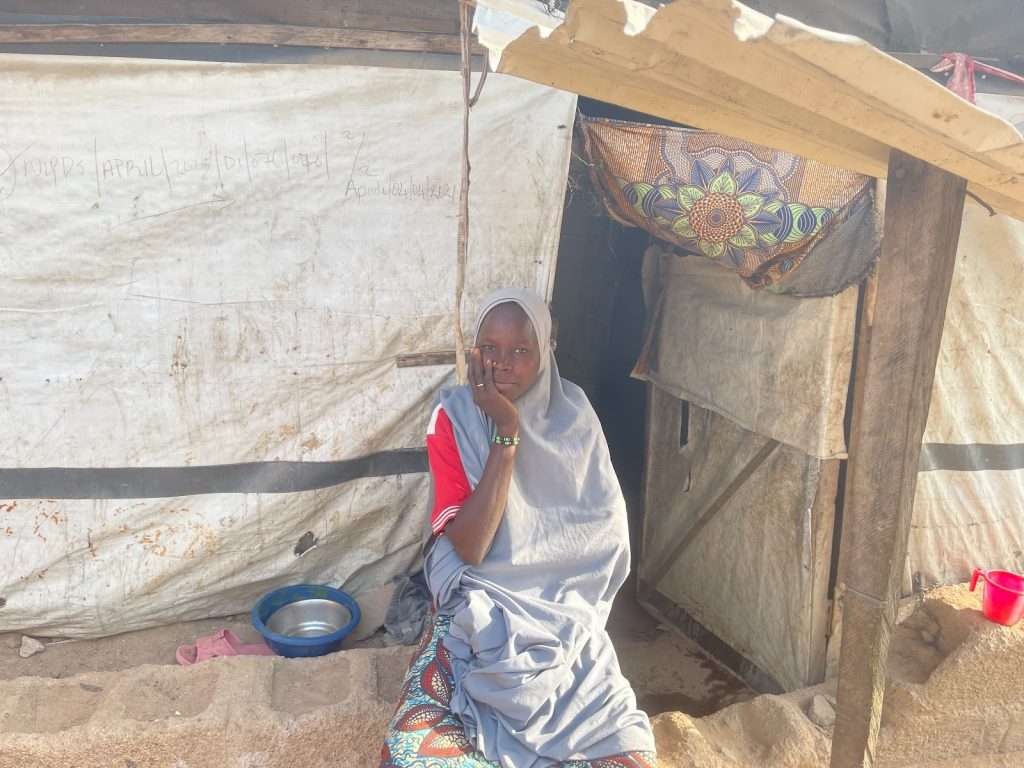 A person in a gray hijab sits outside a makeshift shelter, with a metal bowl on the ground nearby.