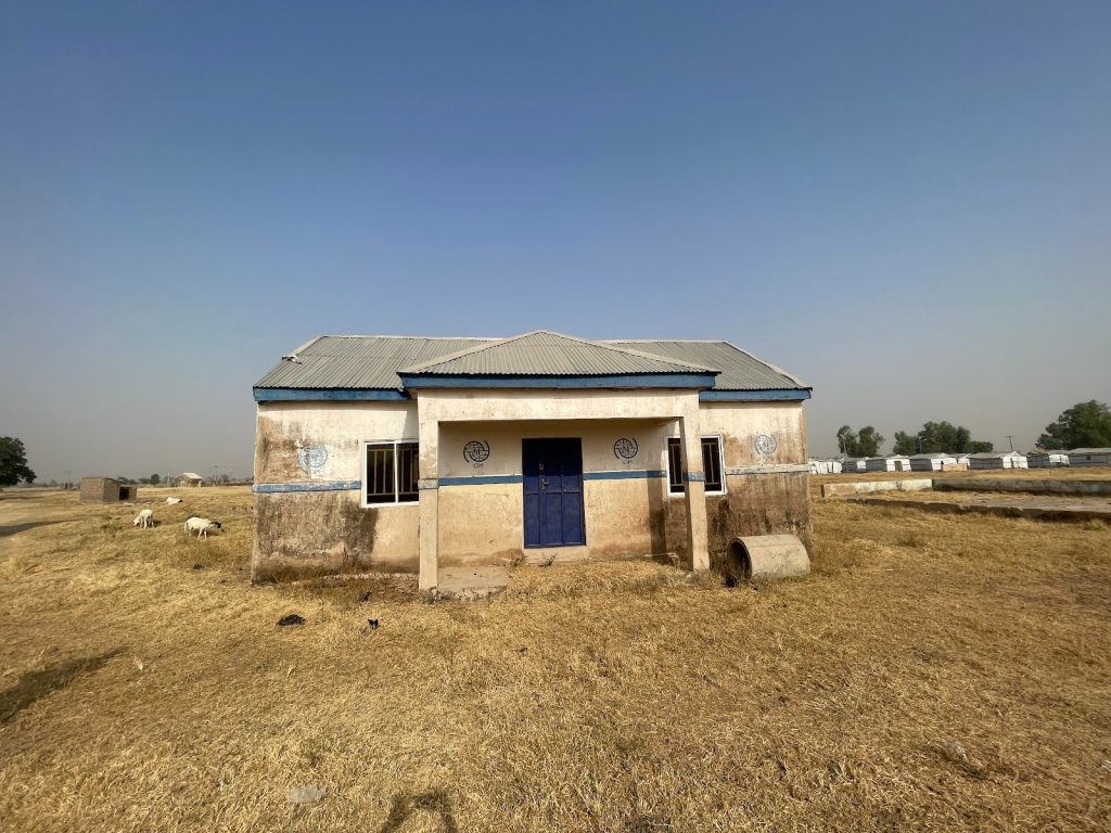 Small, weathered building with a blue door in a dry, grassy field under a clear blue sky. Sheep graze nearby.