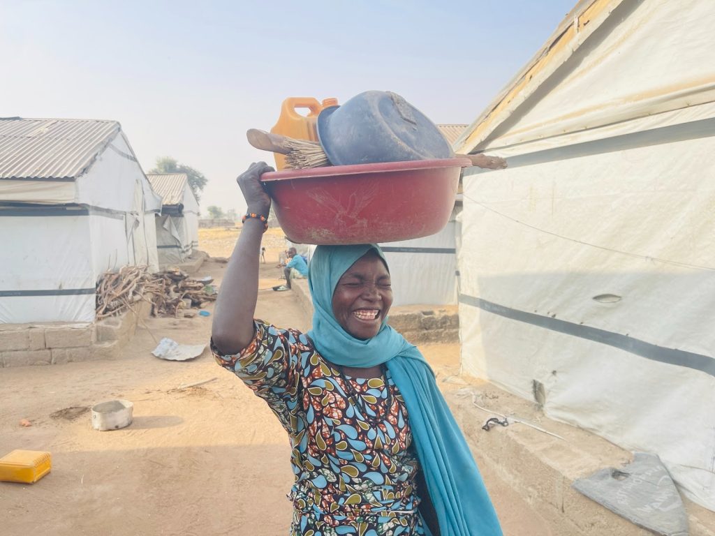 Smiling woman in a colorful dress and headscarf carries a basin with items on her head, walking in front of white tents on a sunny day.