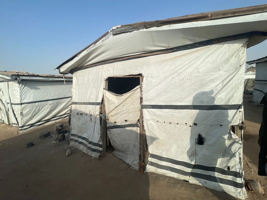 A makeshift shelter with a tarp roof and walls in a dusty area, casting a shadow on its surface.