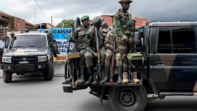 Soldiers in camouflage gear sitting and standing on a military vehicle parked on a street, with another vehicle in the background.