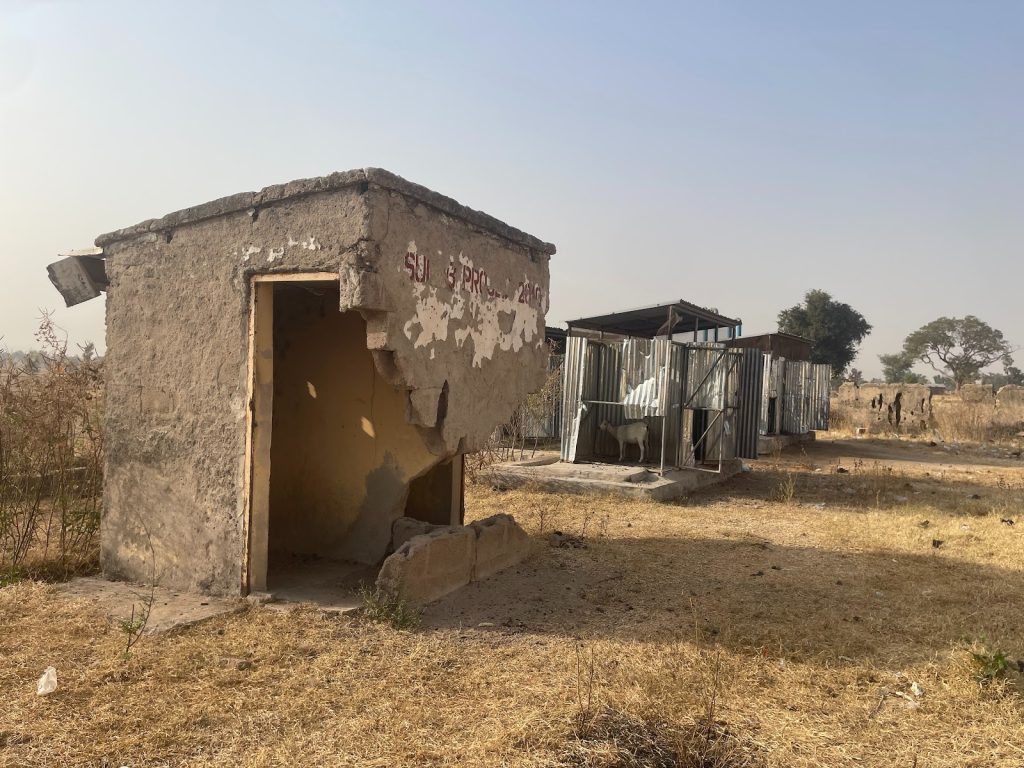 A weathered concrete structure with missing walls stands in a dry field, near makeshift metal shelters with goats.