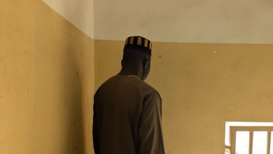Man in a brown outfit and striped hat faces a beige wall, standing next to a window with bars letting in light.