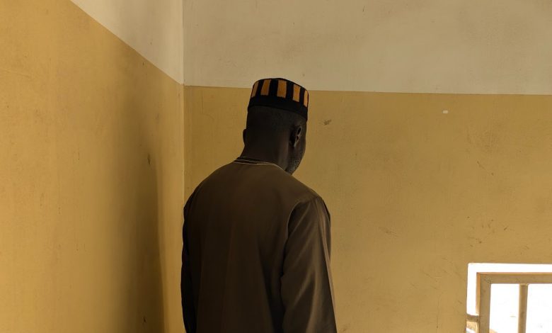 Man in a brown outfit and striped hat faces a beige wall, standing next to a window with bars letting in light.