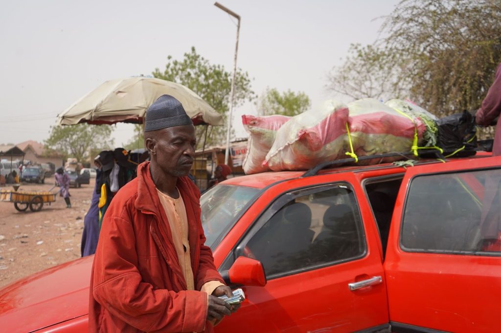 Man in red jacket standing by a red car with sacks on top, holding money. Street scene with a cart and umbrella in the background.