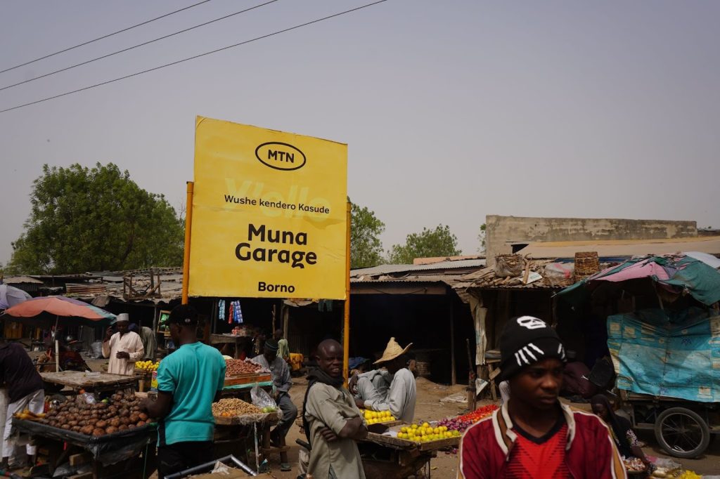 Outdoor market scene with people and colorful produce stalls. A large yellow sign reads "Muna Garage, Borno" with an MTN logo.