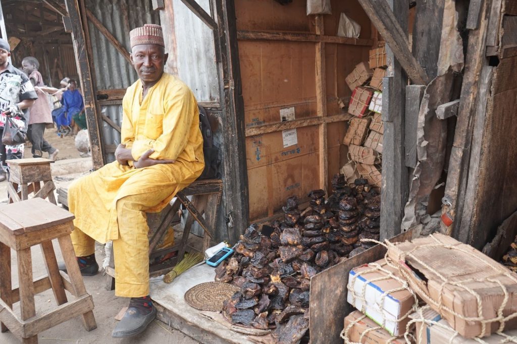 Man in yellow attire sits beside stacks of smoked fish and boxes in a rustic market stall.