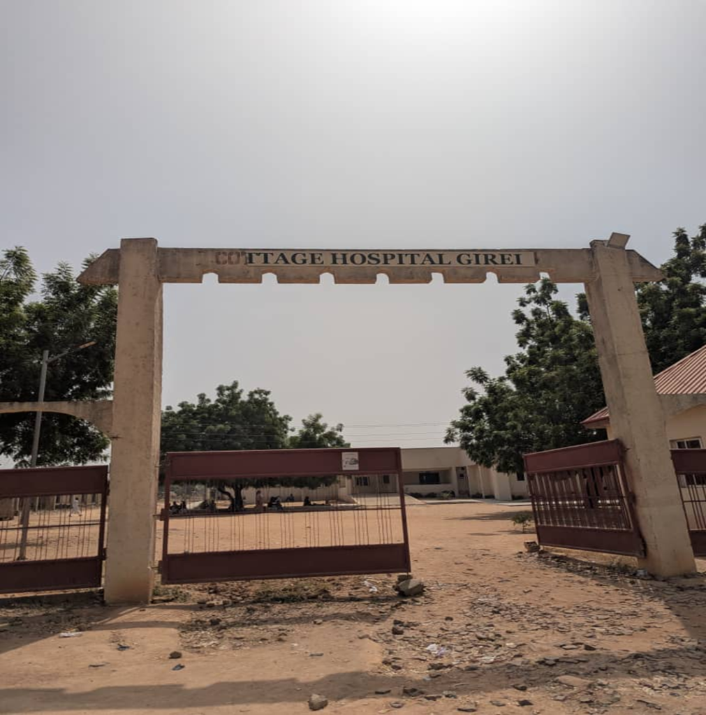 Entrance gate of Cottage Hospital Girei with red metal bars, trees, and a building in the background under a clear sky.