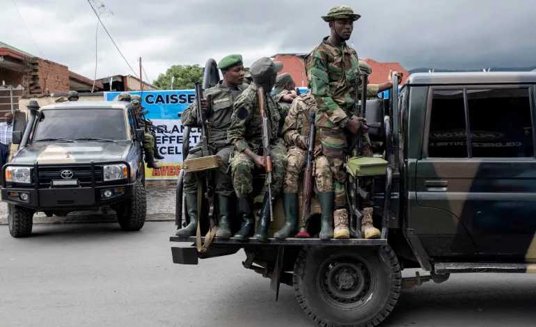 Soldiers in camouflage gear sitting and standing on a military vehicle parked on a street, with another vehicle in the background.