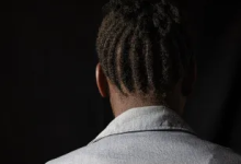Person with dreadlocks, wearing a light-colored shirt, seen from the back against a dark background.