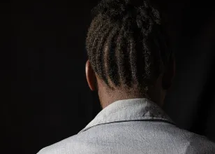 Person with dreadlocks, wearing a light-colored shirt, seen from the back against a dark background.