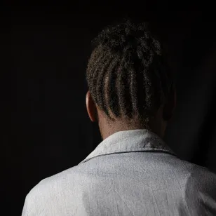 Person with dreadlocks, wearing a light-colored shirt, seen from the back against a dark background.